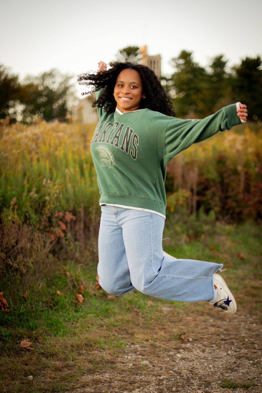 Person in green sweatshirt and jeans jumps playfully outdoors in autumn setting.