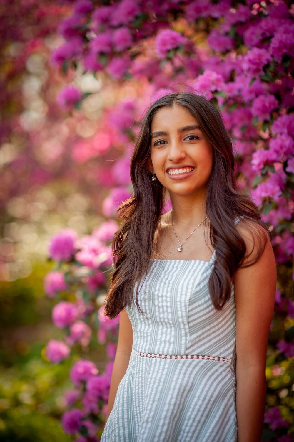 Young woman in a light blue dress smiling in front of vibrant pink flowering bushes outdoors.