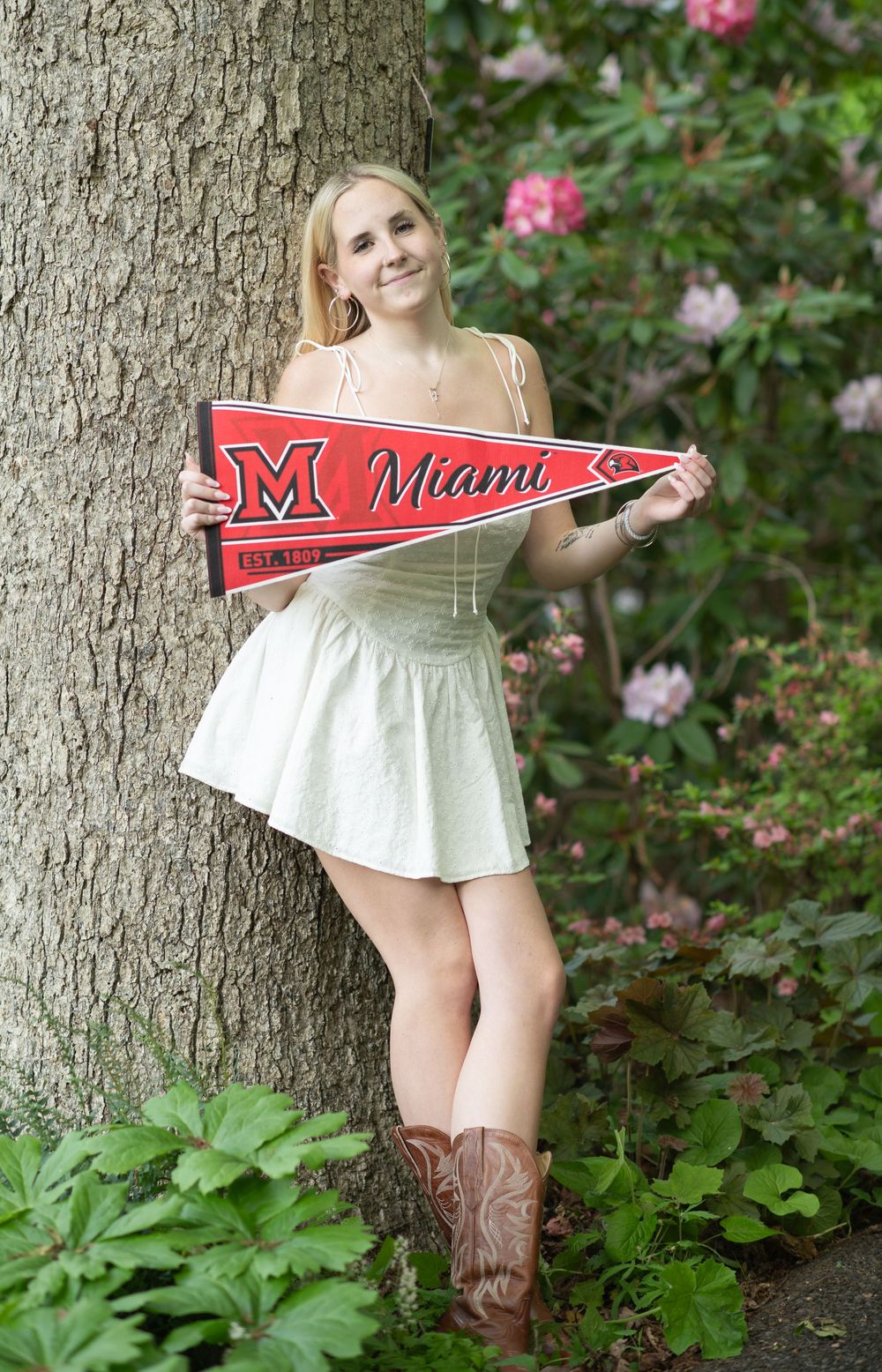 Smiling blonde woman in white dress and cowboy boots holding a Miami University red pennant flag outdoors.