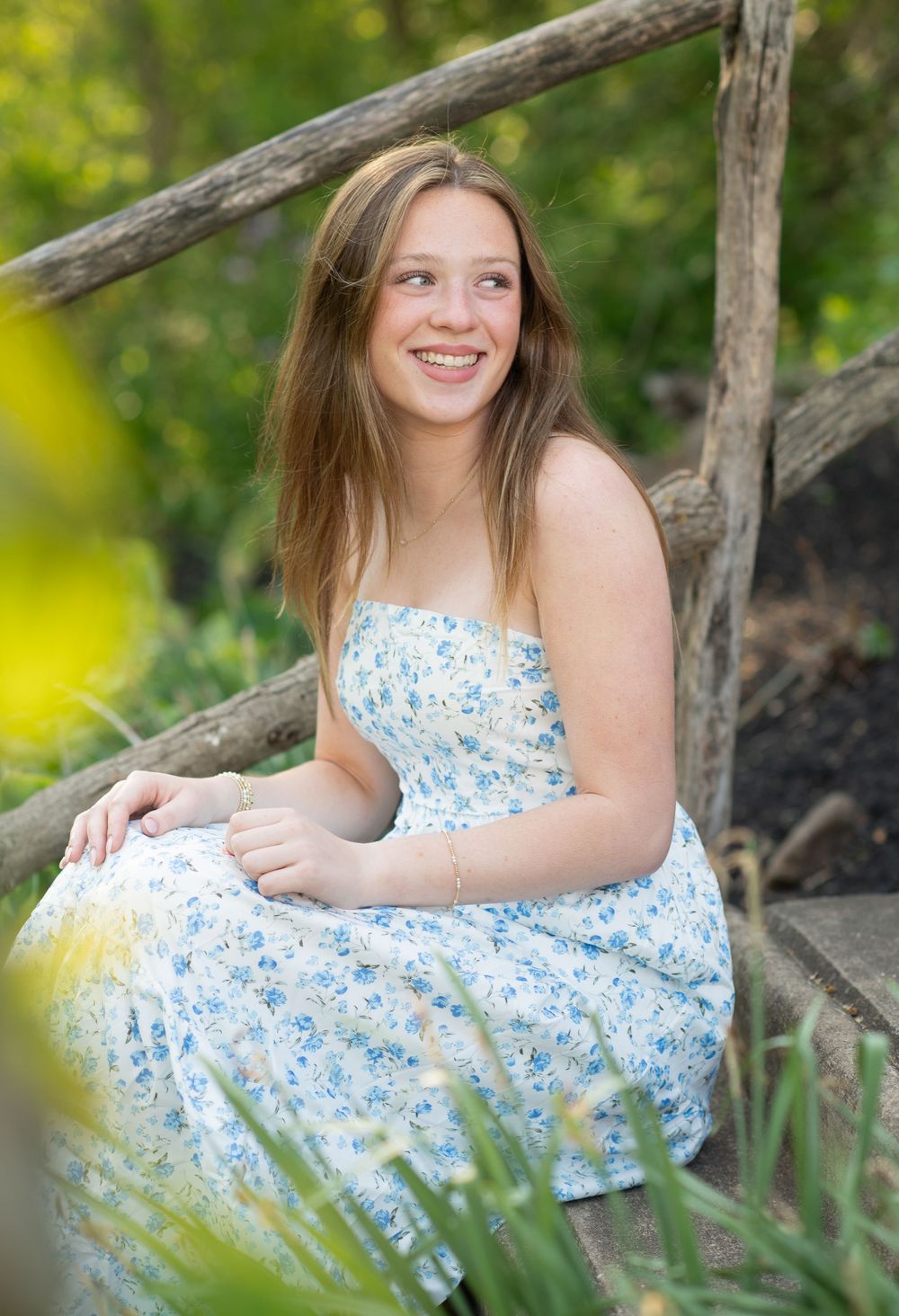 Young woman in floral sundress smiling outdoors on rustic wooden steps surrounded by lush green garden foliage.