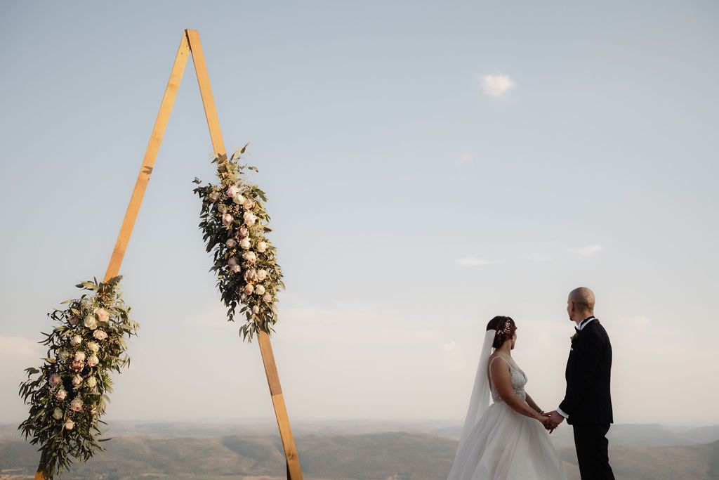 A couple in wedding attire holds hands during sunset ceremony beneath a triangular floral arch on a hilltop.