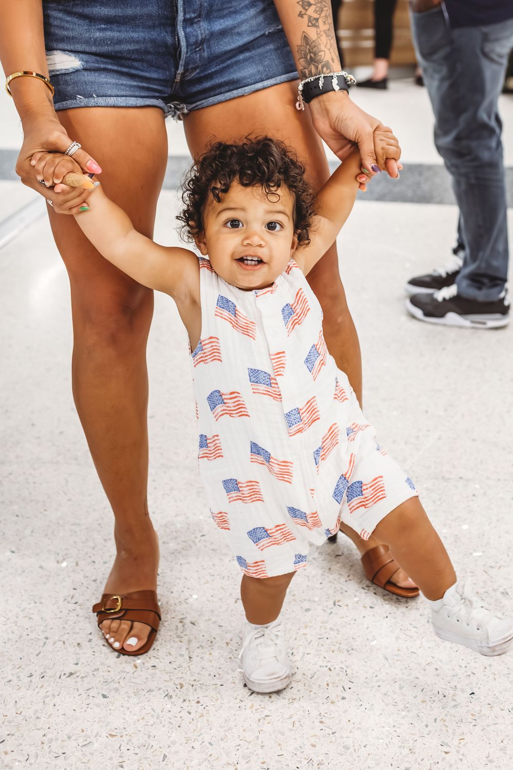 Small child wearing American flag outfit learning to walk on white tile floor in Hamden, CT.
