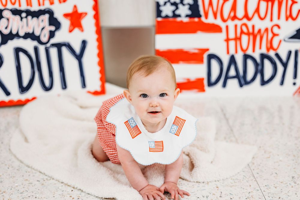 Baby in white onesie lying on floor next to patriotic welcome home signs in Hamden, CT.