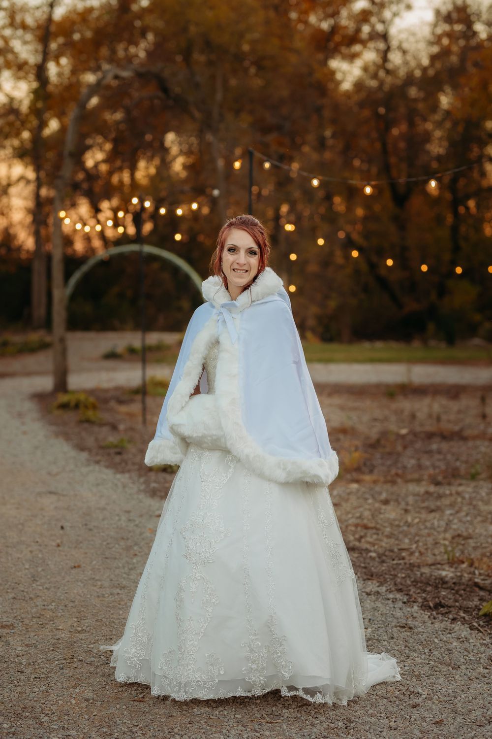 A bride in a white lace wedding dress poses outdoors during golden hour with autumn trees in the background.