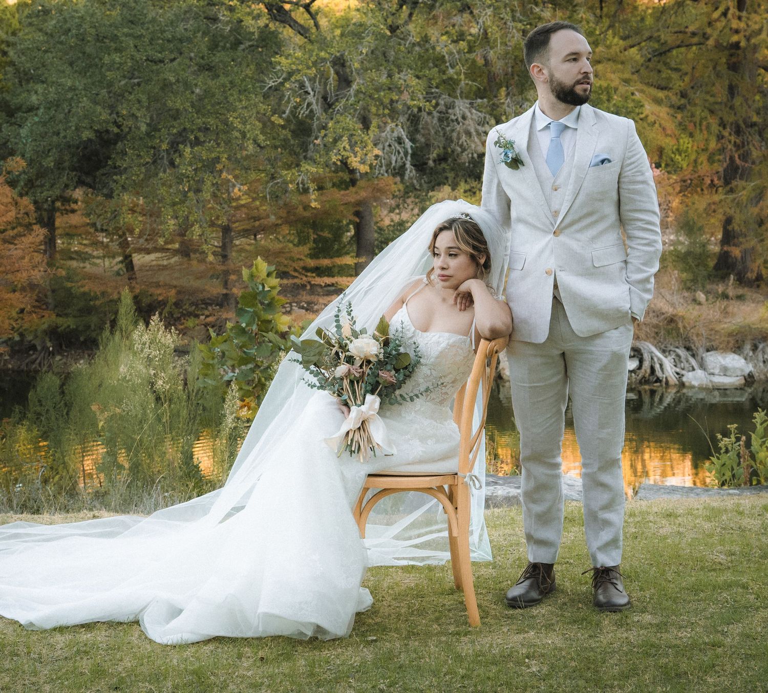 A romantic wedding couple sits together on vintage chairs by a serene lake during golden hour in an autumn setting.