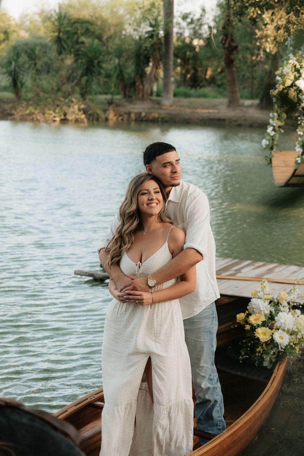 Couple in white outfits embrace romantically on a wooden boat by a serene lake with lush greenery.