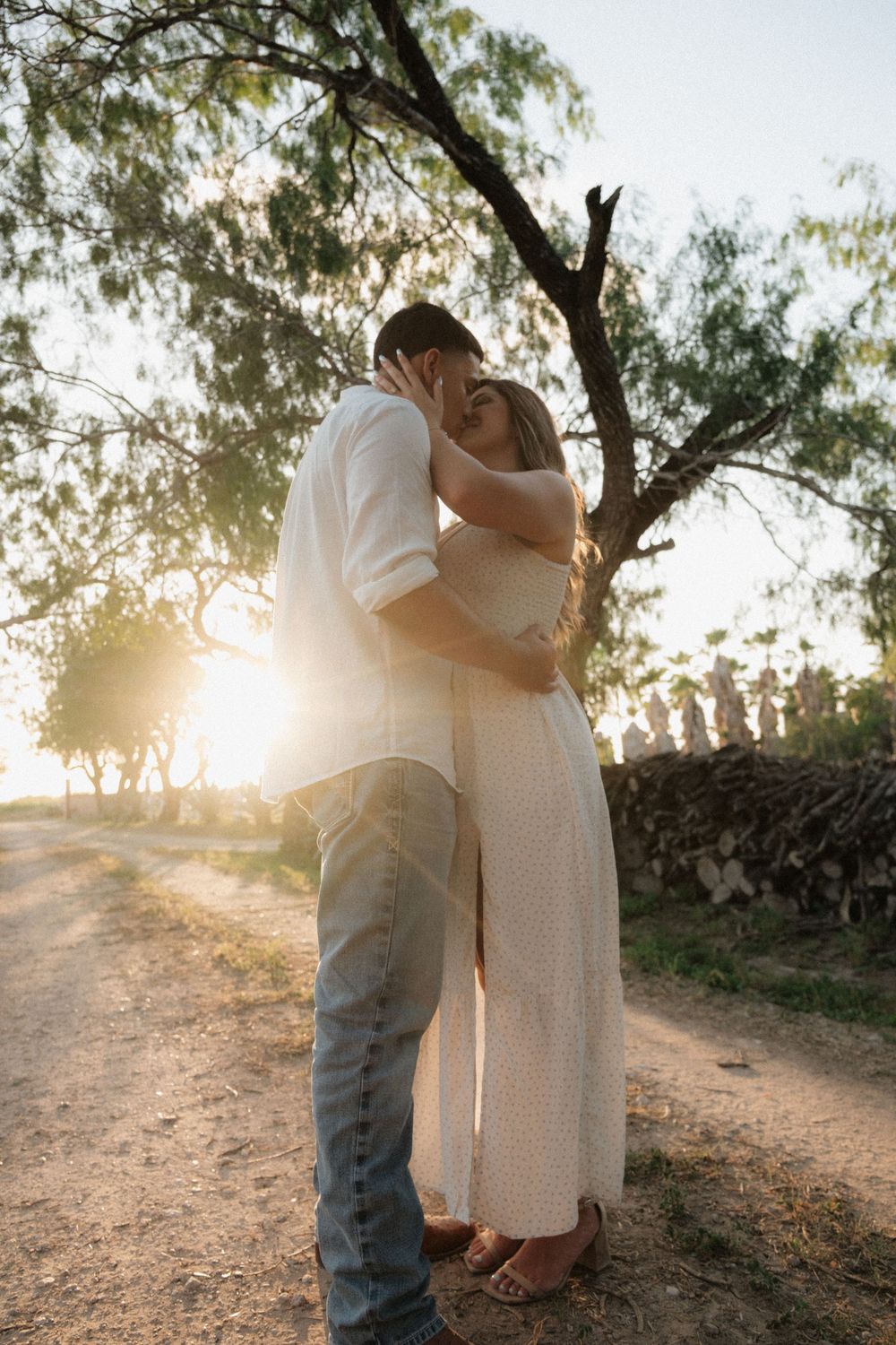 Couple embracing and kissing at golden hour sunset on a dirt path surrounded by trees.