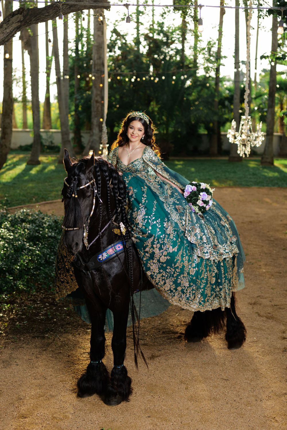 A stunning teal and silver quinceañera dress flows elegantly beside a majestic black horse in a wooded setting.