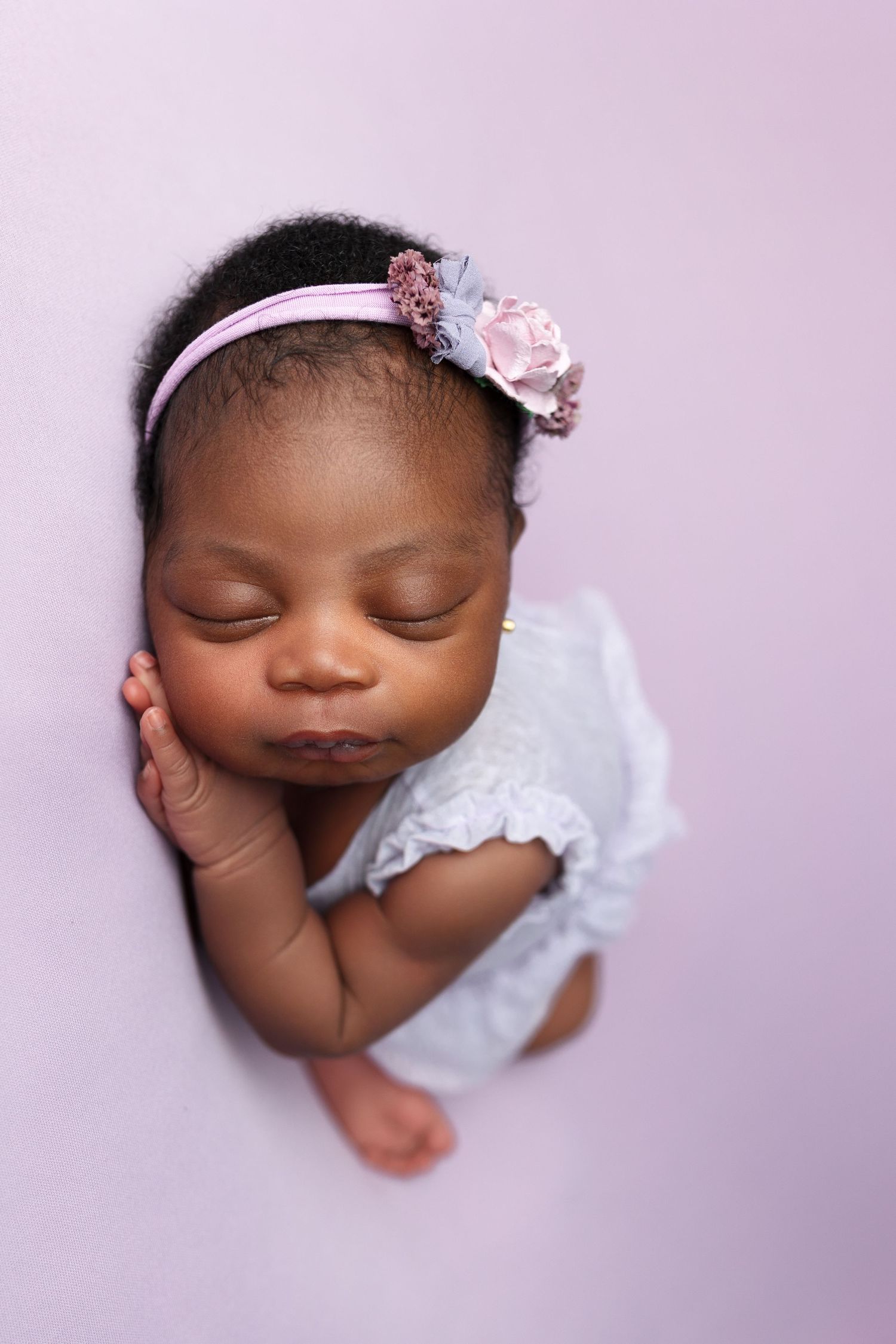 Sleeping newborn in white lace dress and floral headband on purple background.