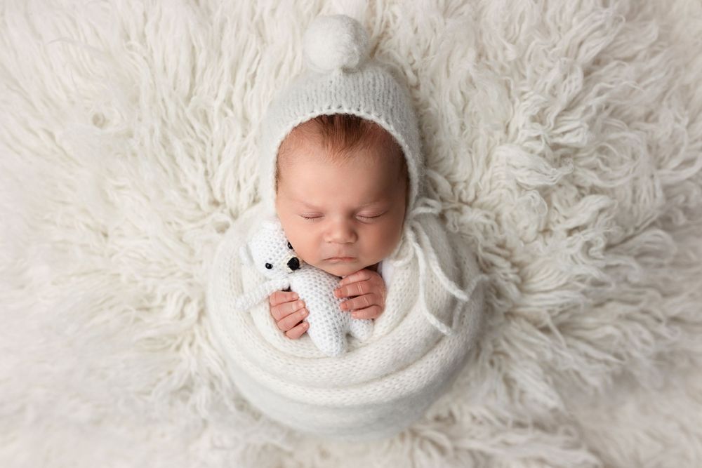 A sleeping newborn baby swaddled in white lies on soft white fur blanket in a professional photography setting.