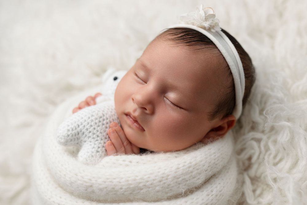 Sleeping newborn wrapped in white knit blanket on soft white textured backdrop.