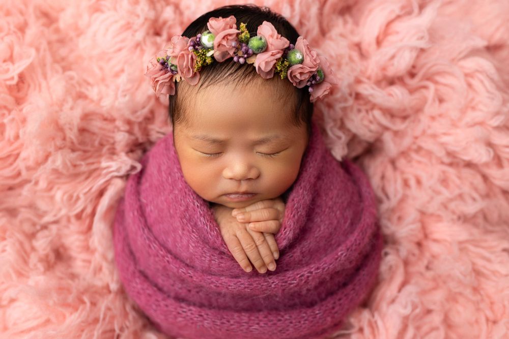 Sleeping baby wearing floral crown wrapped in pink blanket on pink textured backdrop.