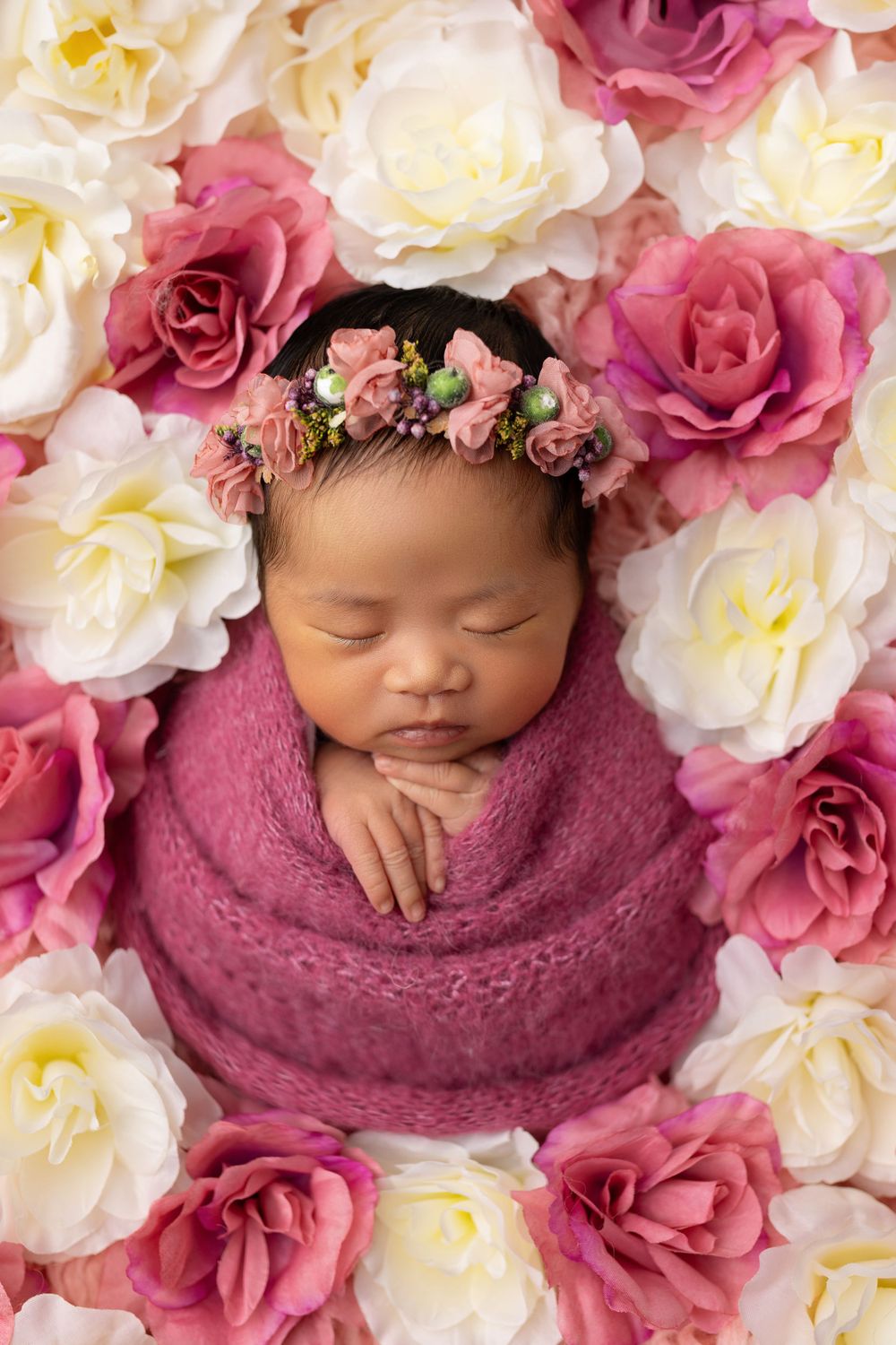 Peaceful sleeping newborn surrounded by pink and white roses wearing delicate floral crown.