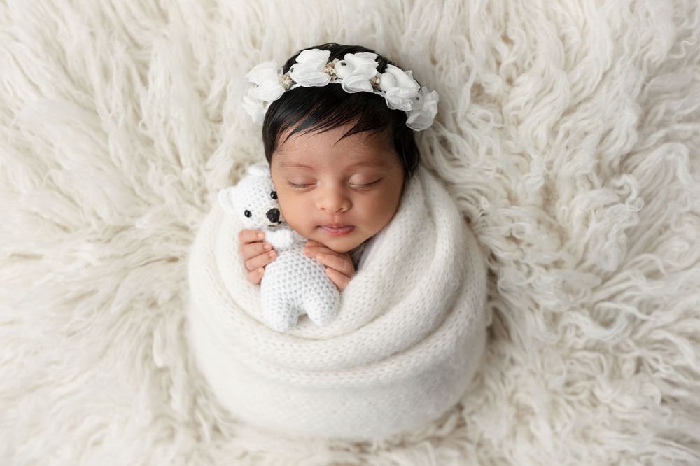 Newborn baby sleeping peacefully on soft white fur blanket with delicate flower headband.
