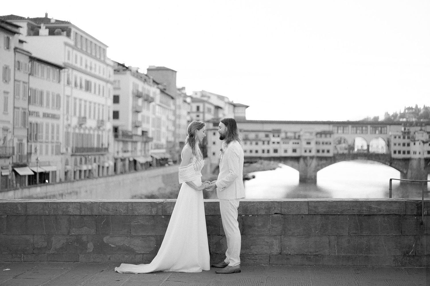Couple in their wedding dresses facing each other and holding their hands while exchanging vows at Ponte Vecchio, Florence, Italy
