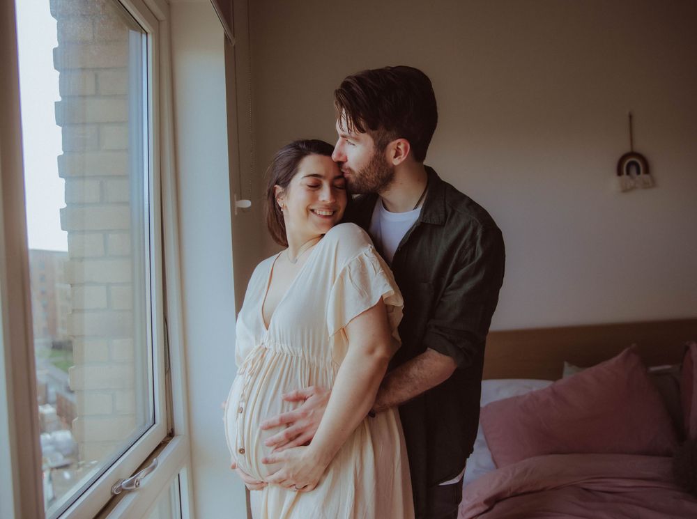 Expecting couple shares tender moment near sunlit window in cozy bedroom with pink pillows and minimal decor.