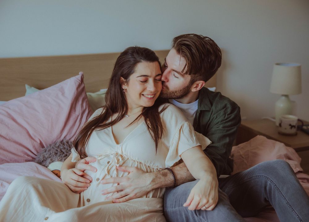 A couple shares an intimate moment while cuddling together on a bed with pink bedding and a neutral backdrop.