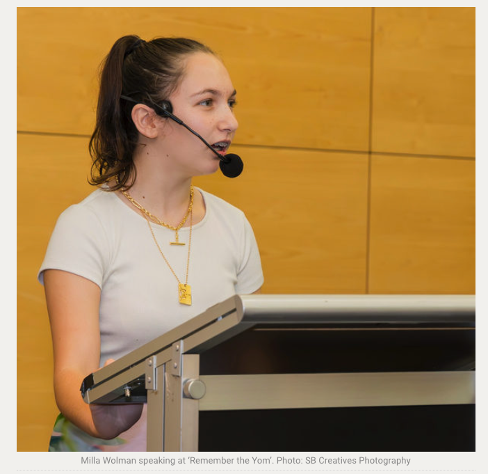 A speaker wearing a headset microphone presents at a podium against a yellow wall.