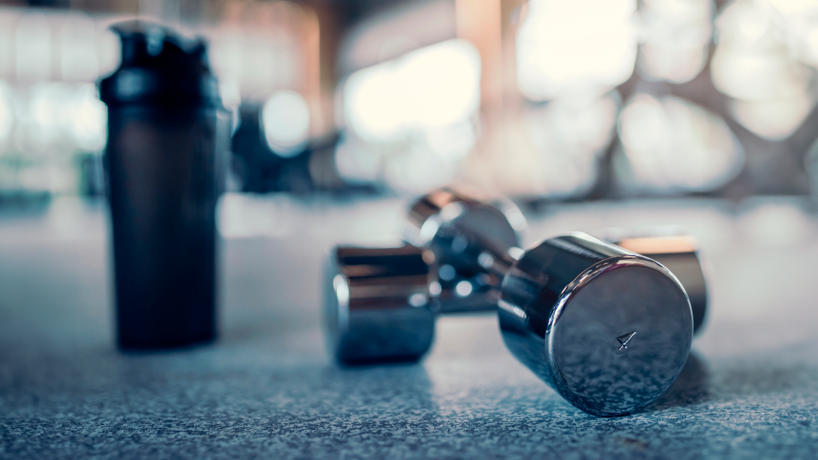 Close-up of dumbbells and protein shaker on gym floor.