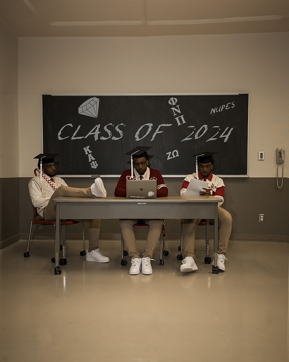 Three students sit at a desk with a chalkboard showing "Class of 2024" behind them.