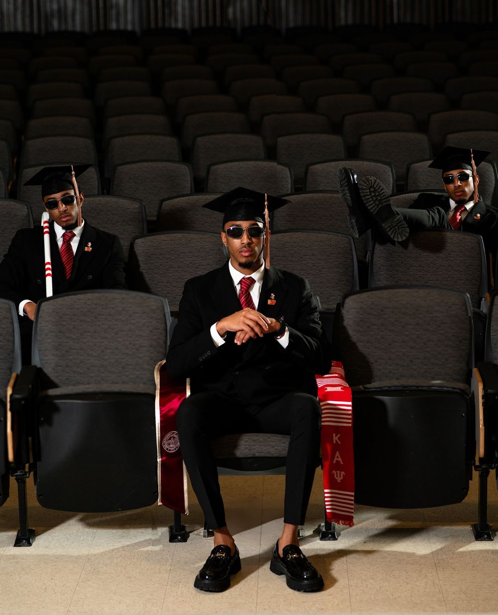 Three graduates in sunglasses and black suits pose in an auditorium with red graduation stoles and mortarboards.