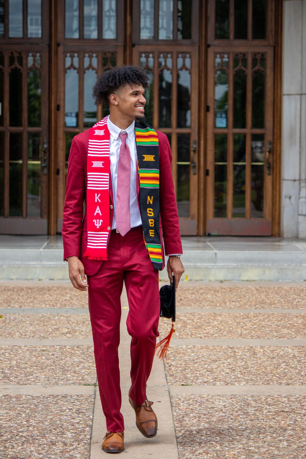 Stylish maroon suit with cultural stoles and brown dress shoes walking on ornate stone pavement.
