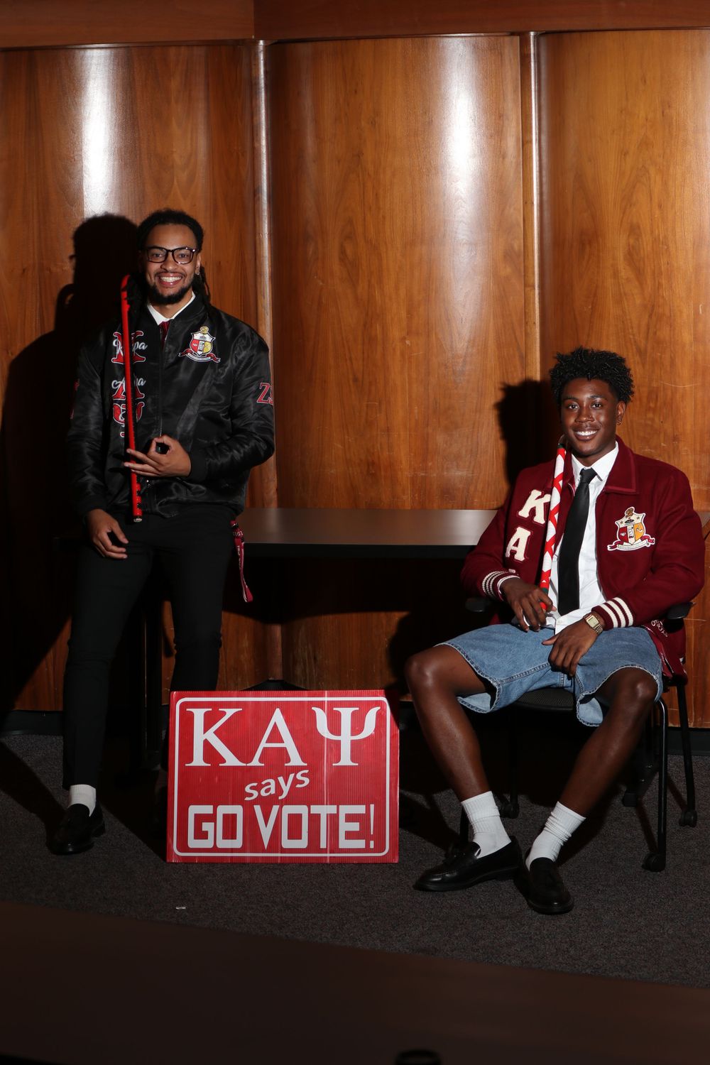 Fraternity brothers posing with 'Go Vote' sign in wood-paneled room wearing varsity jackets.