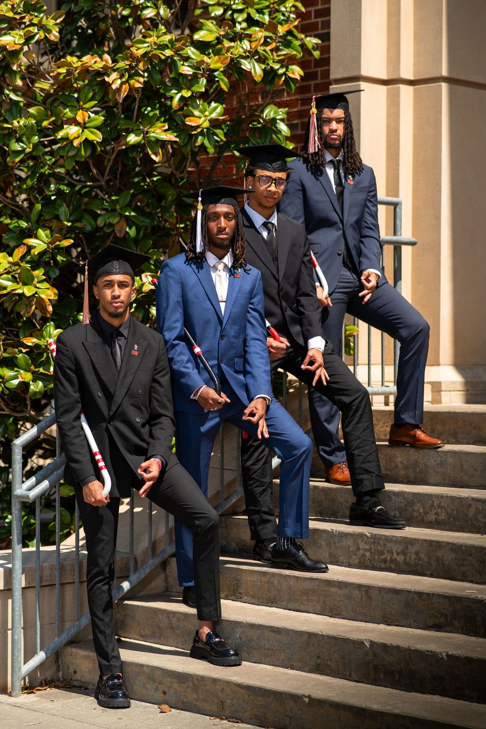 Graduates in formal suits and caps stand on stone steps near a magnolia tree during a college graduation ceremony.