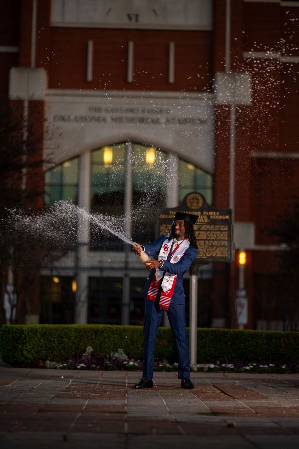 A graduate stands in front of a building at night holding a champagne bottle while spraying it in celebration.