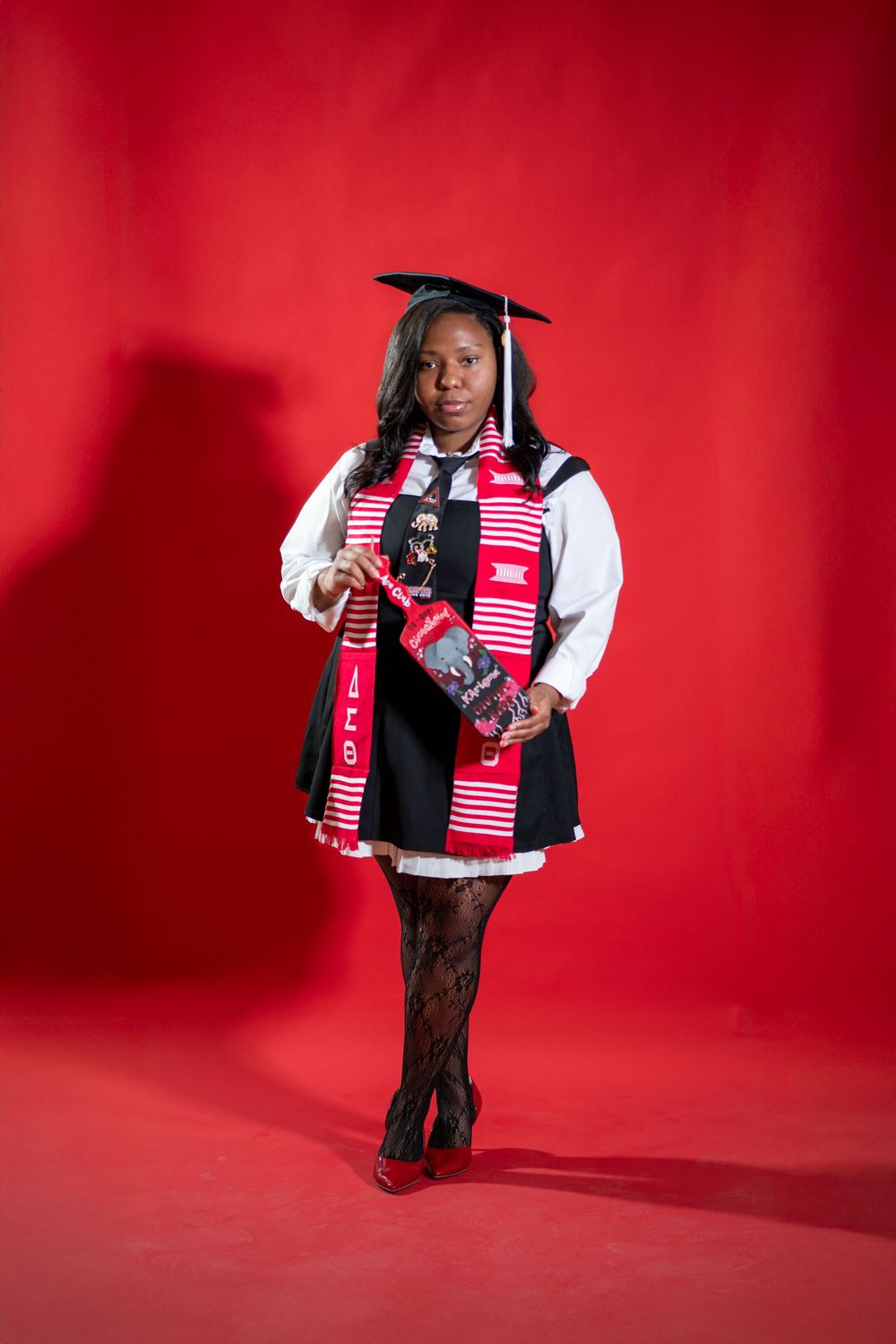 Graduate in black and white academic regalia with red and white stole poses against vibrant red backdrop for portrait.