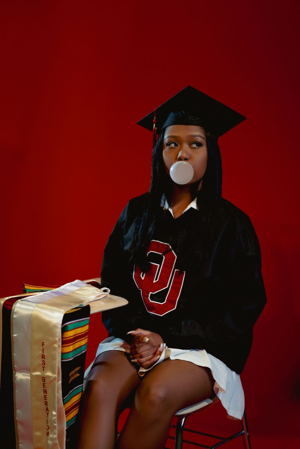 Graduate in black cap and gown with OU logo blows bubble gum while posing against red backdrop with suitcase.