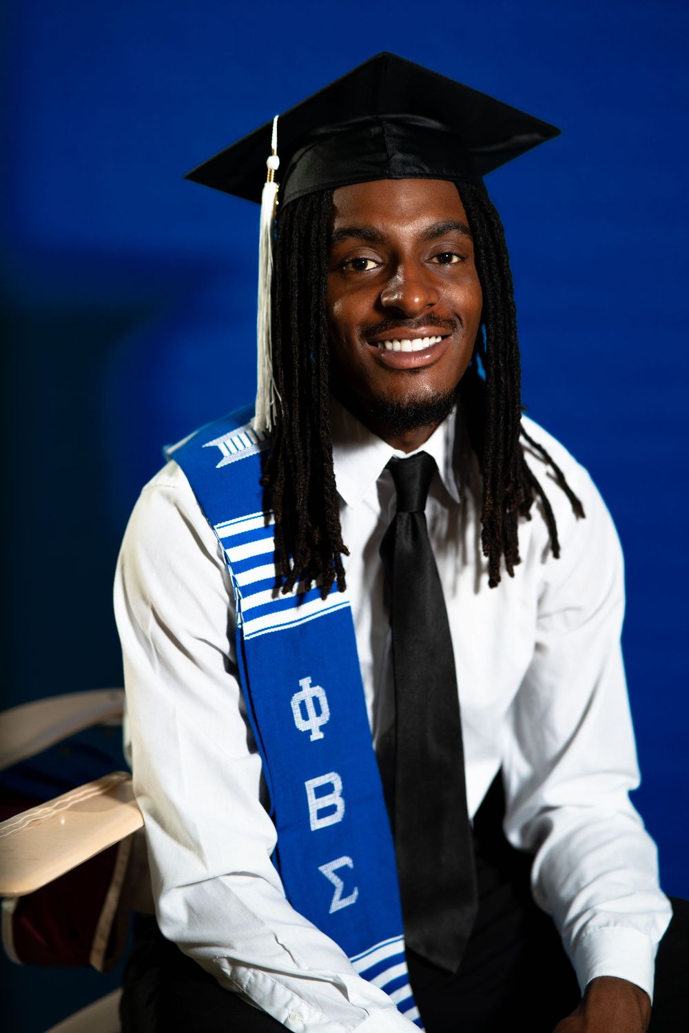 Graduate in cap and gown wearing black tie and white shirt with blue academic sash during graduation photo session.