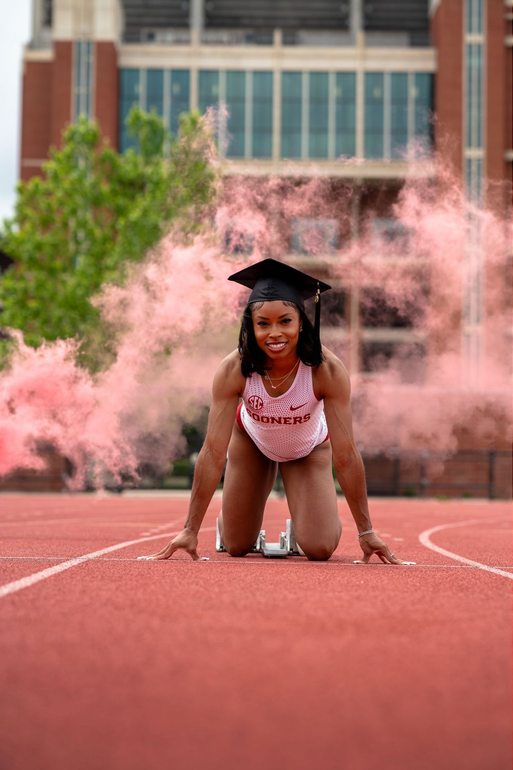 Graduate in starting position on running track with pink smoke in background at university track field.
