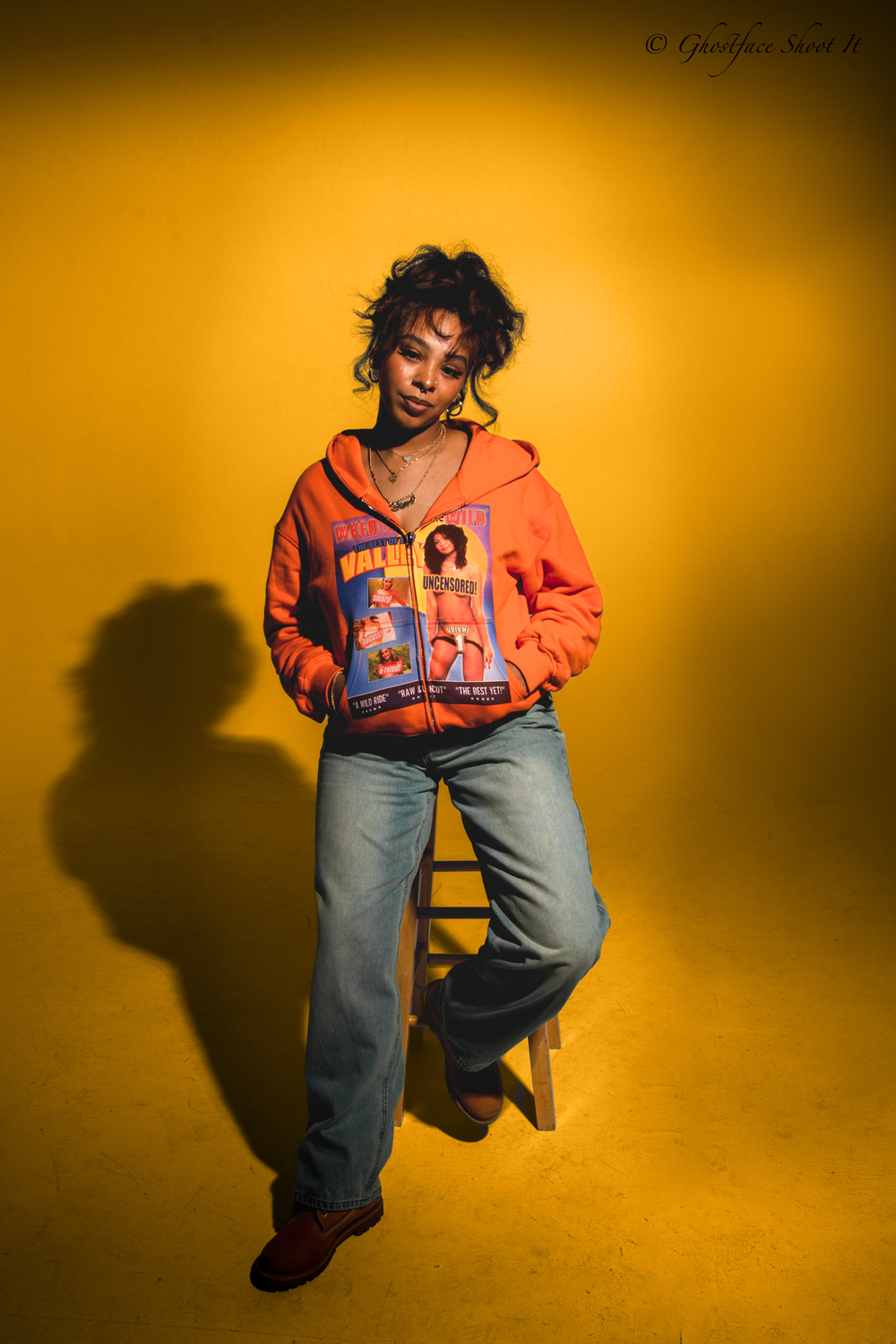 Person in orange hoodie poses against yellow backdrop with dramatic lighting.