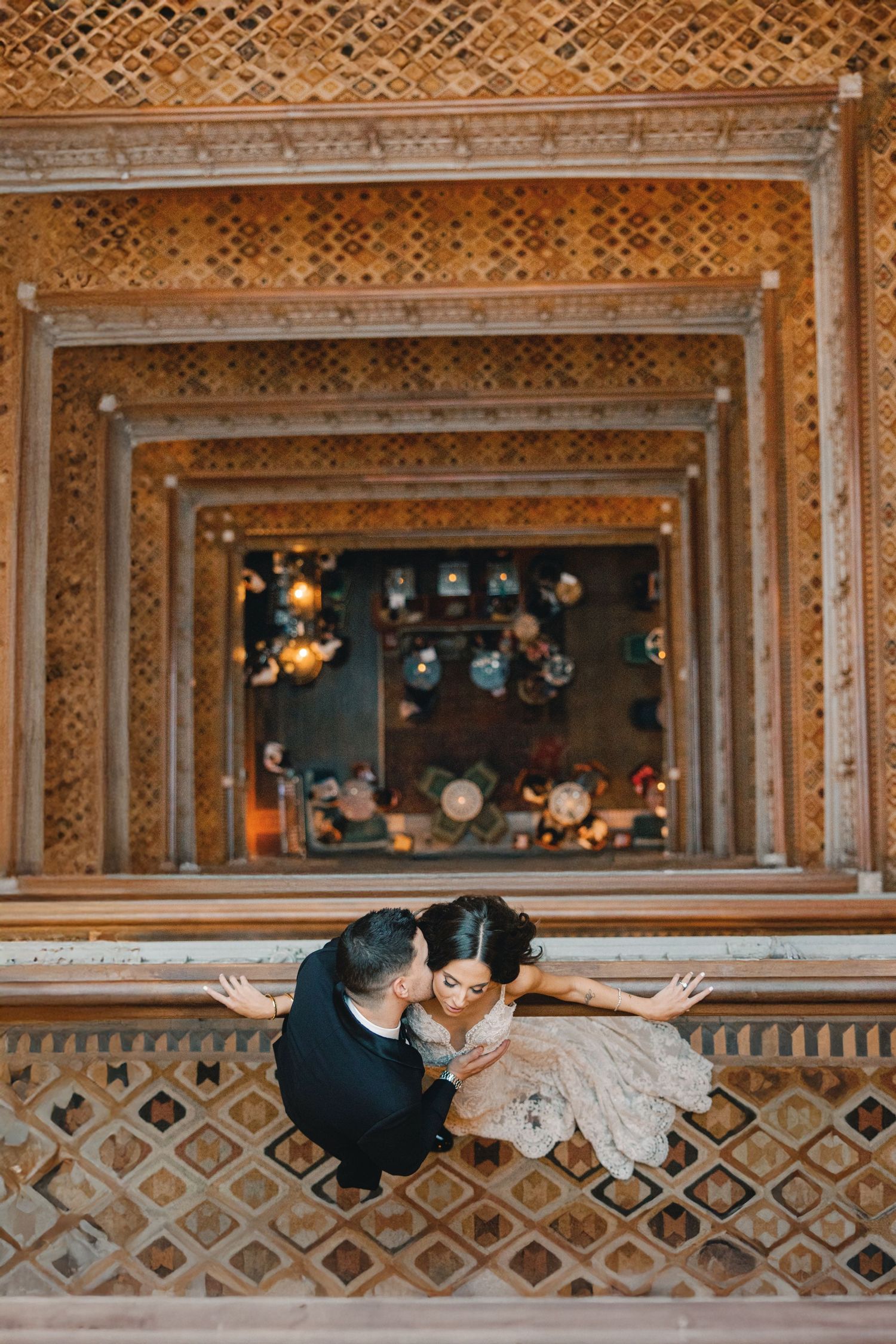Aerial view of wedding couple sharing a kiss on ornate balcony with decorative carved ceiling above.
