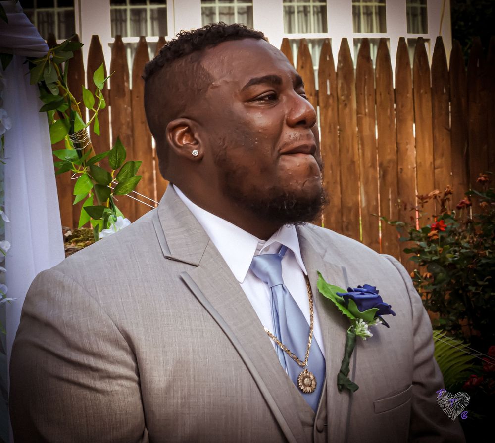 A formal portrait outdoors featuring a grey suit with boutonniere against a wooden fence backdrop.