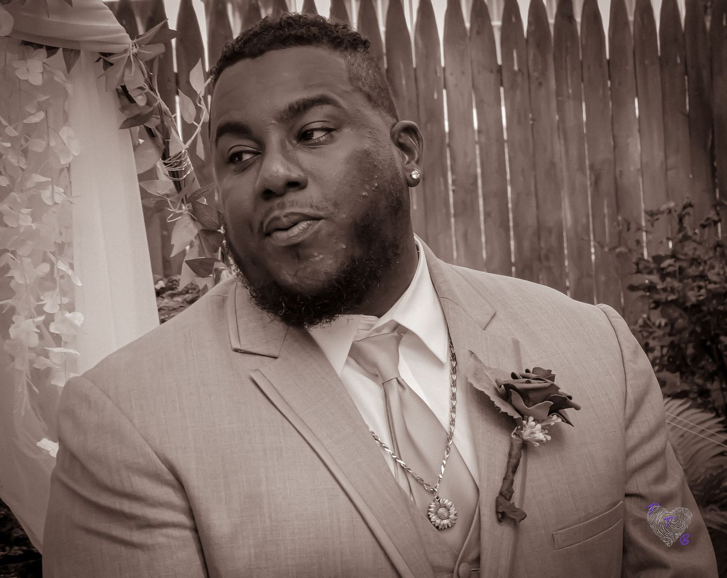 A formal portrait in black and white featuring a grey suit and boutonniere against a wooden fence.
