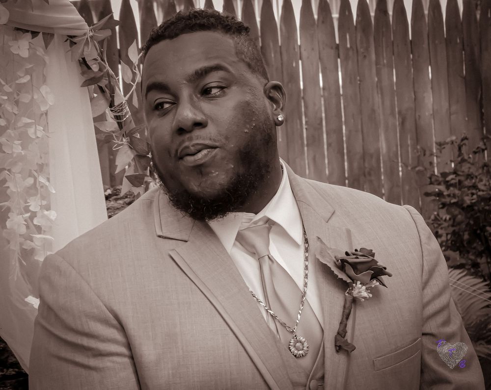 A formal portrait in black and white featuring a grey suit and boutonniere against a wooden fence.