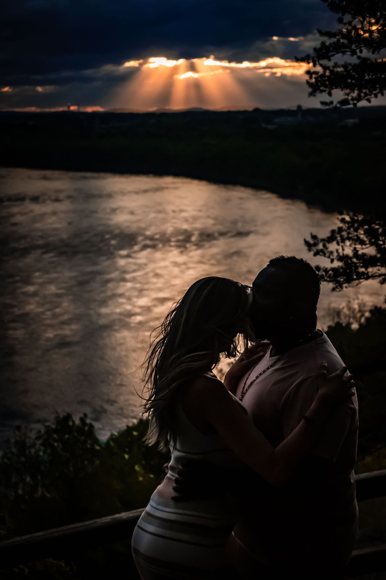 Silhouetted couple embracing at sunset with dramatic sunbeams breaking through clouds over a river.