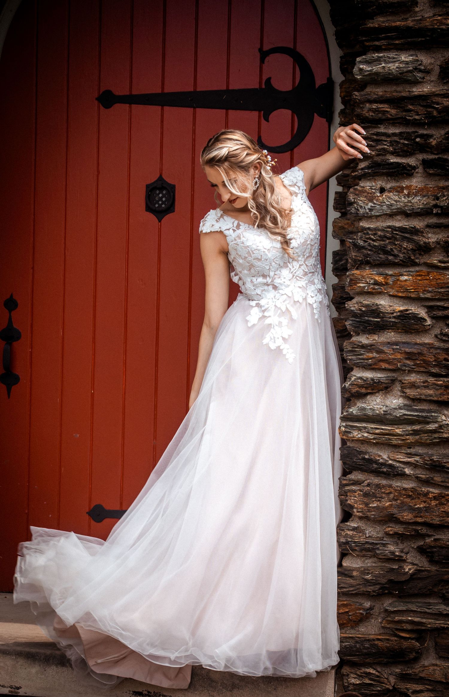 Bride in lace wedding gown poses against red door and stone wall.