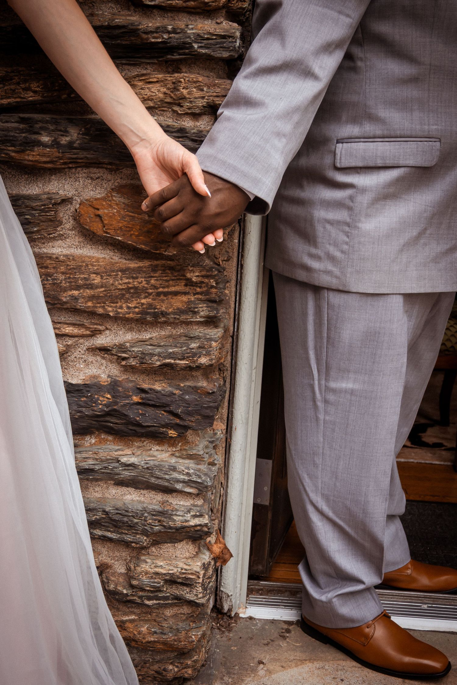 A couple in wedding attire joins hands against a rustic log cabin wall.