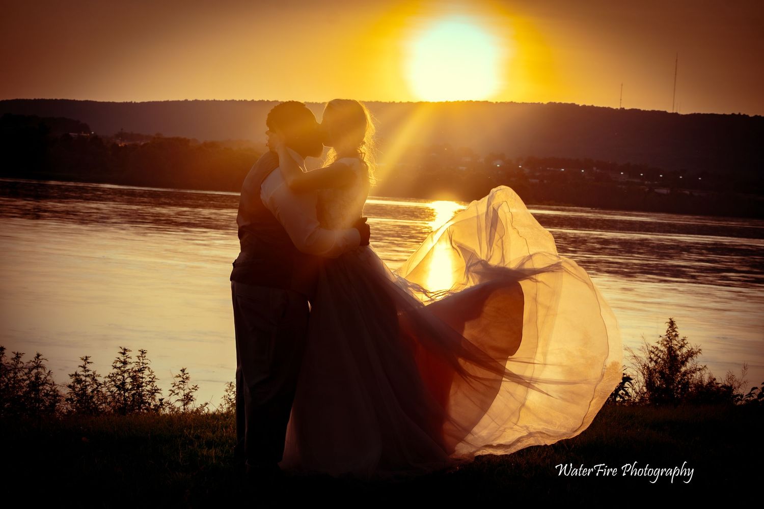 Silhouette of couple at sunset with floating wedding dress veil by a lake.