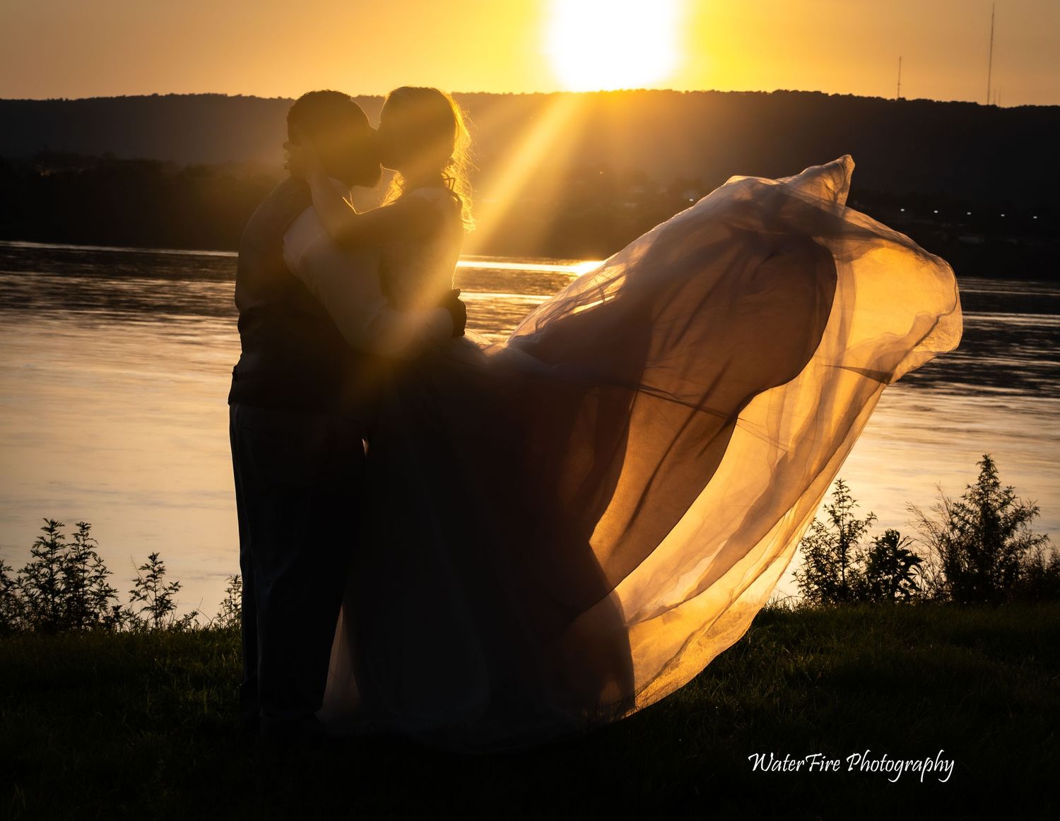 Wedding dress veil flows in breeze as couple stands against golden sunset over water.
