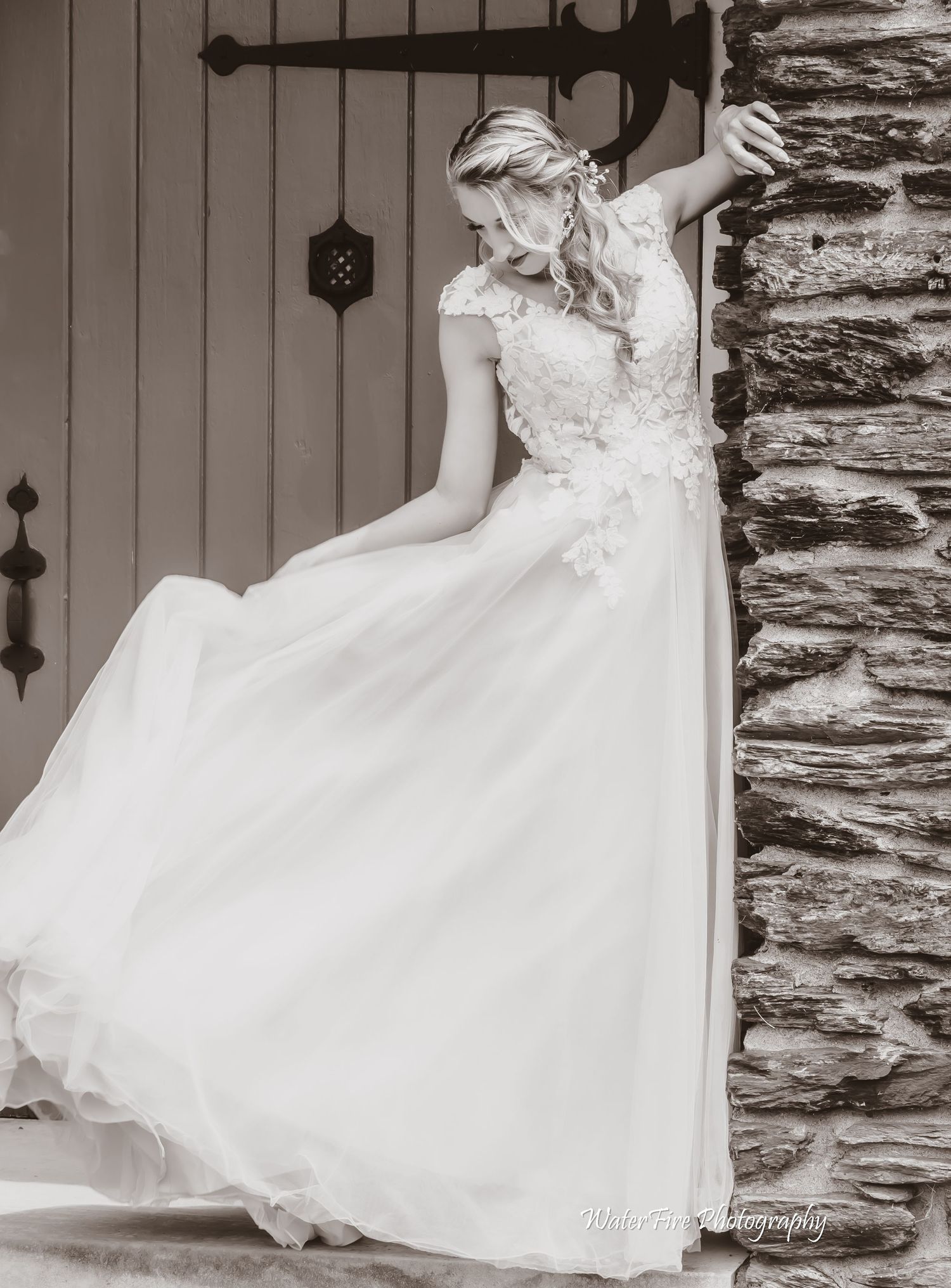 Bride in flowing white lace gown poses against stone wall in black and white.