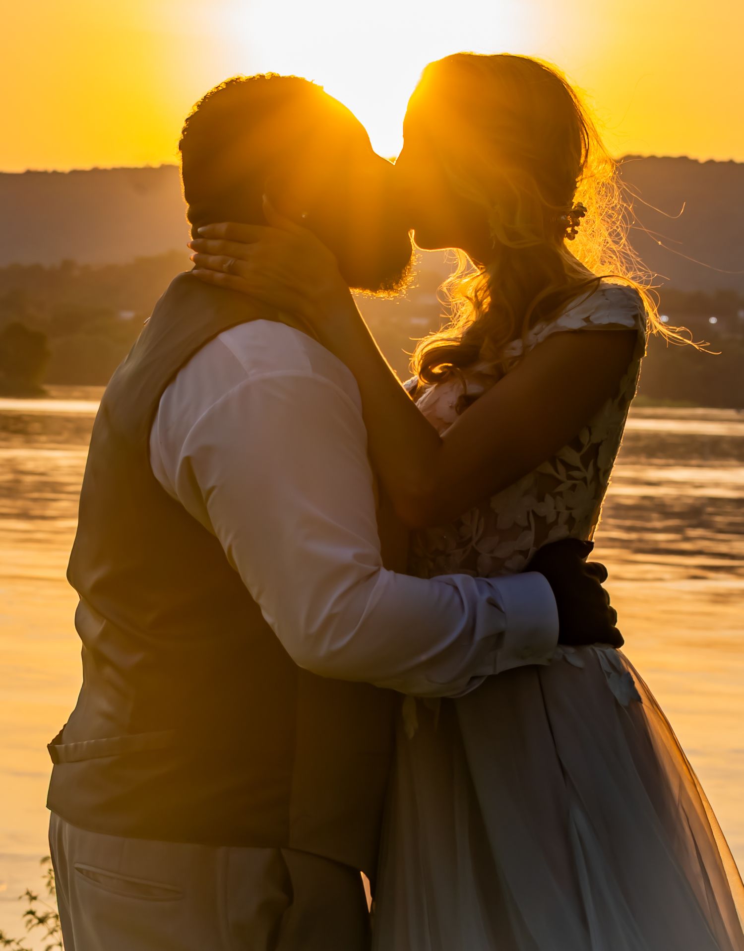 Backlit silhouette of couple kissing during golden hour by lake.