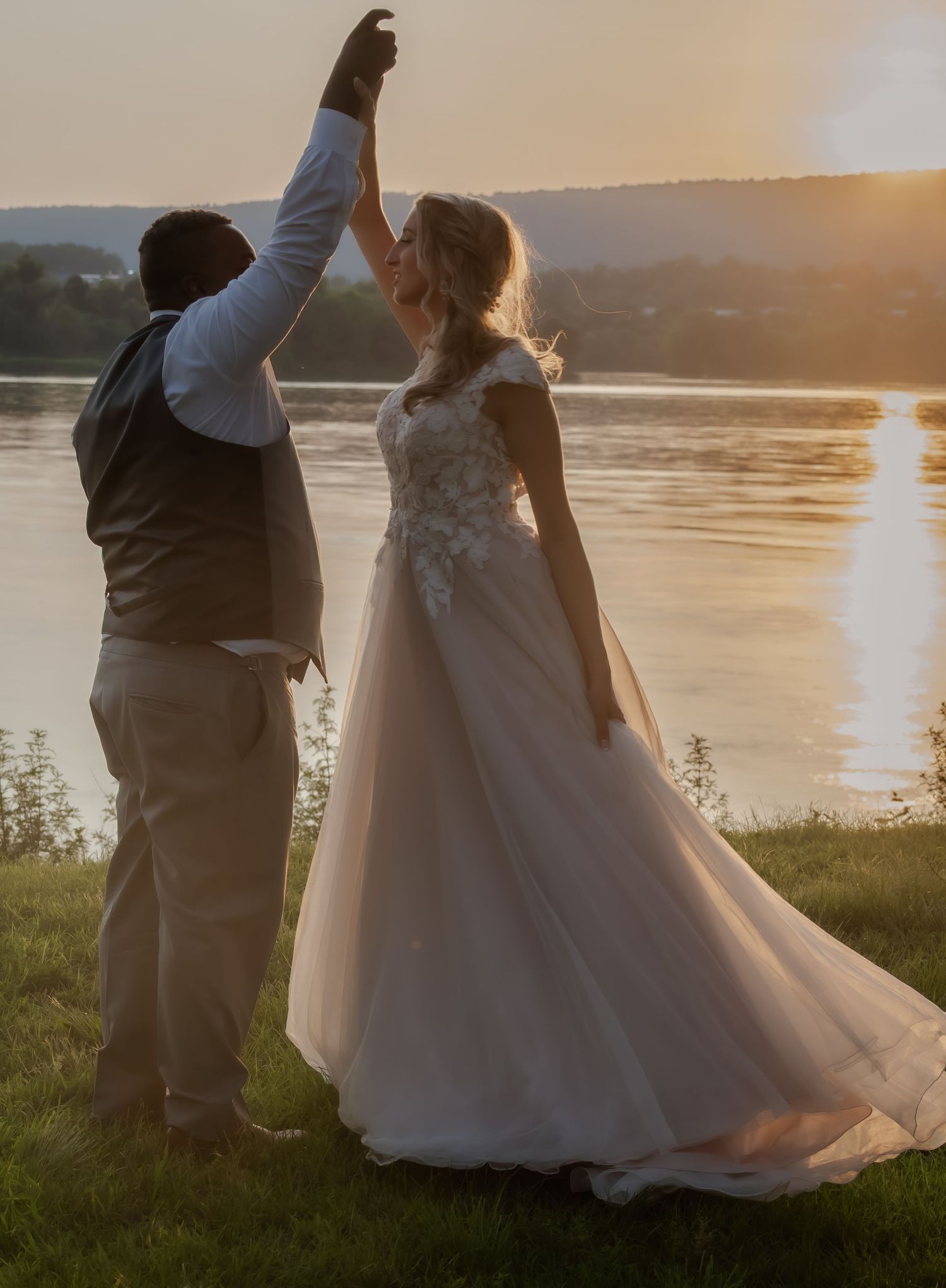 A wedding couple celebrates with raised hands against a scenic lakeside sunset backdrop.