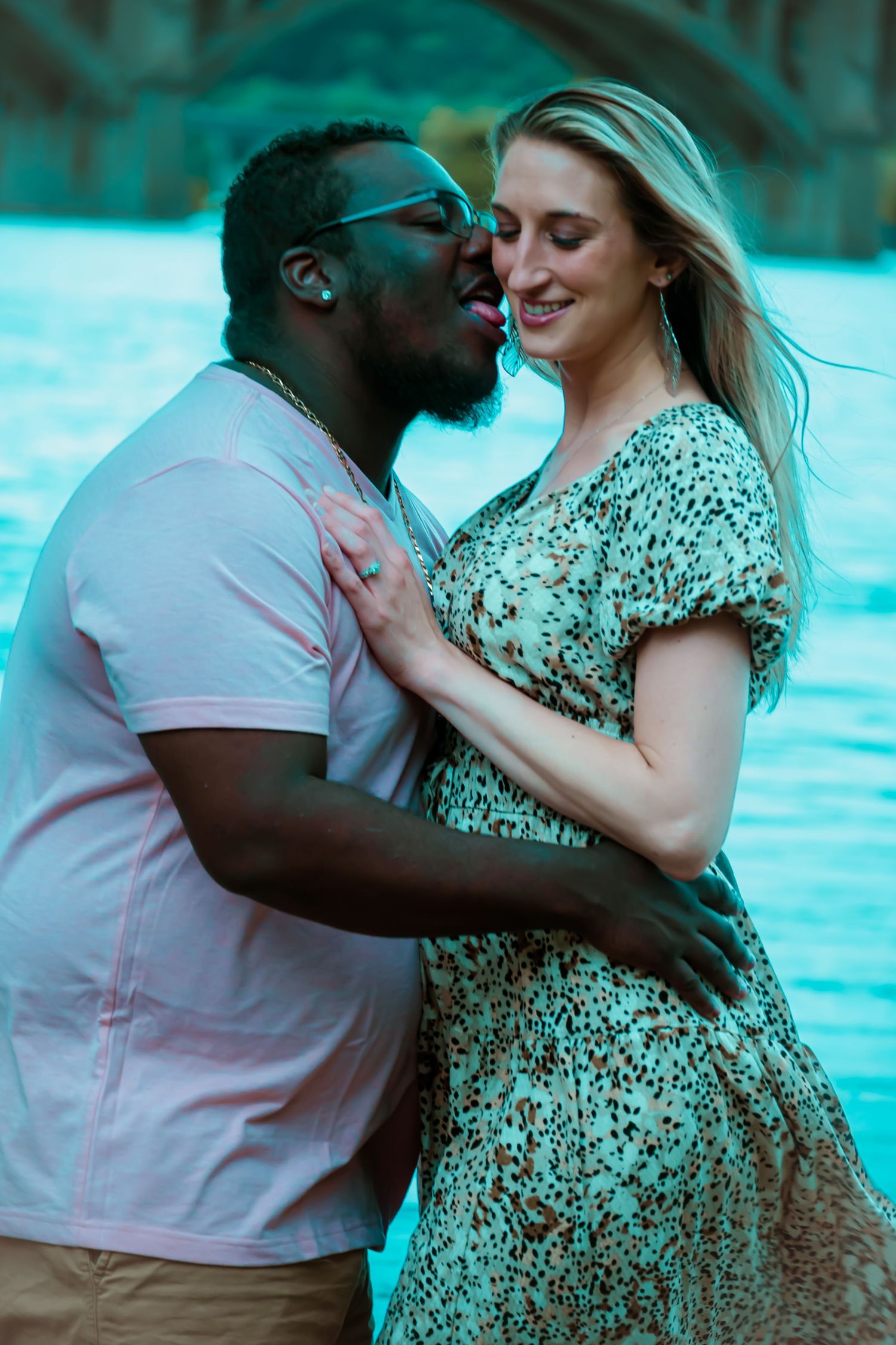 A couple shares an intimate moment by a waterfront, with the woman wearing a patterned dress and the man in a pink polo.