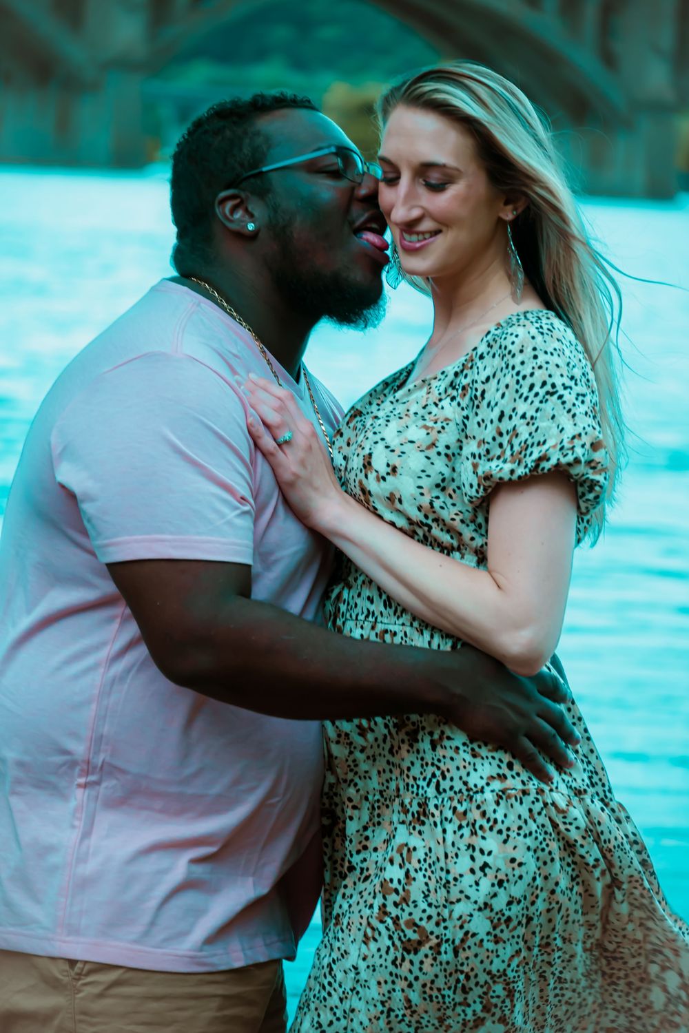 A couple shares an intimate moment by a waterfront, with the woman wearing a patterned dress and the man in a pink polo.