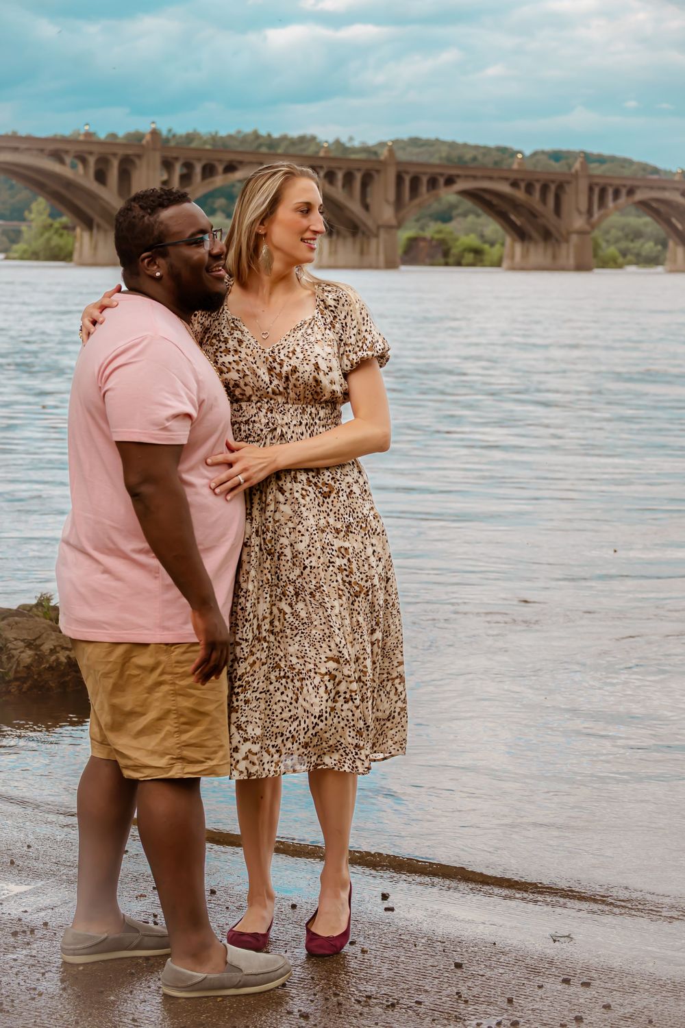 A couple stands together near a bridge over water, wearing summer casual attire.