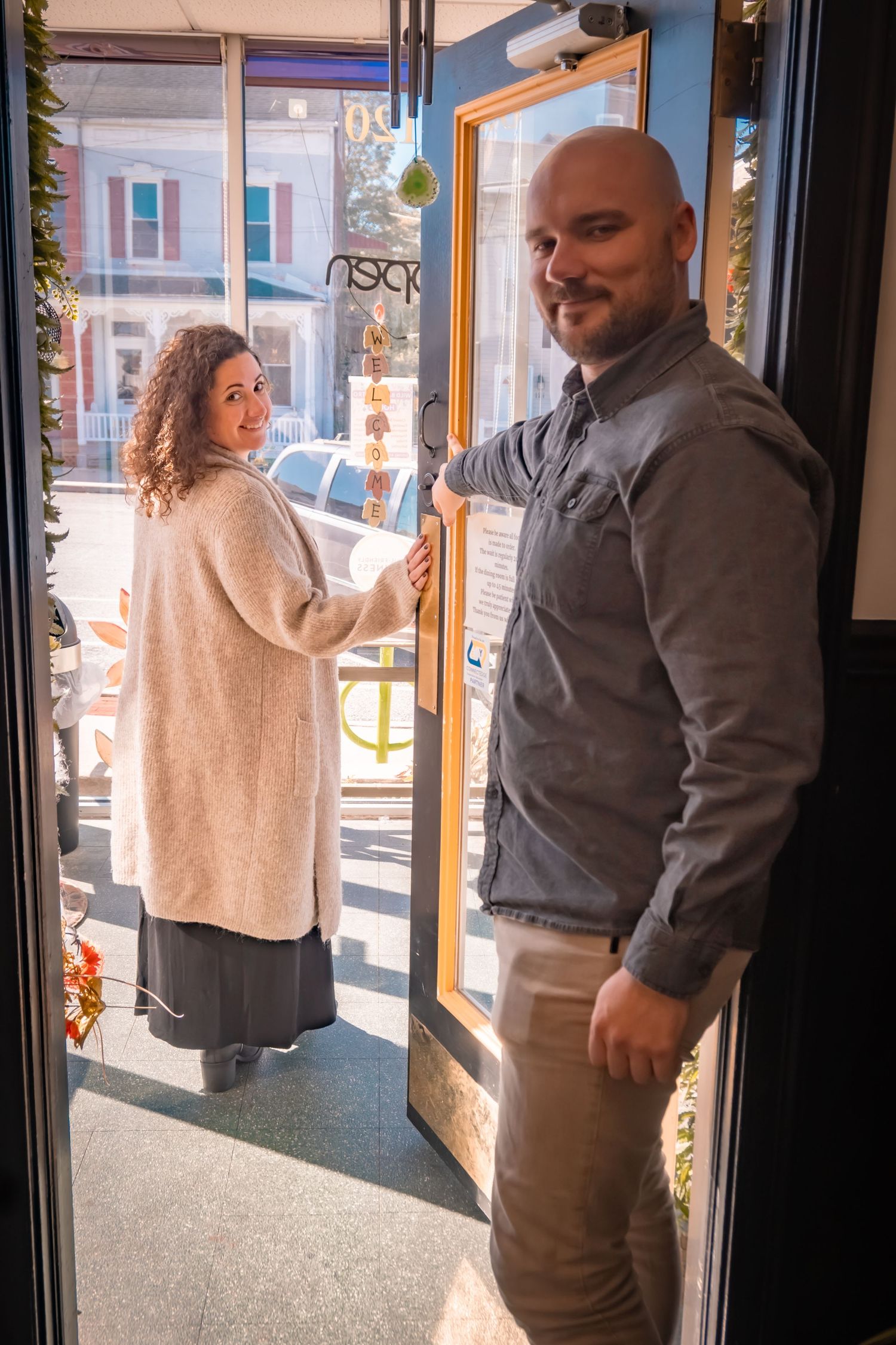 A bald man holds open a shop door for a curly-haired woman in a cream cardigan, both smiling warmly.